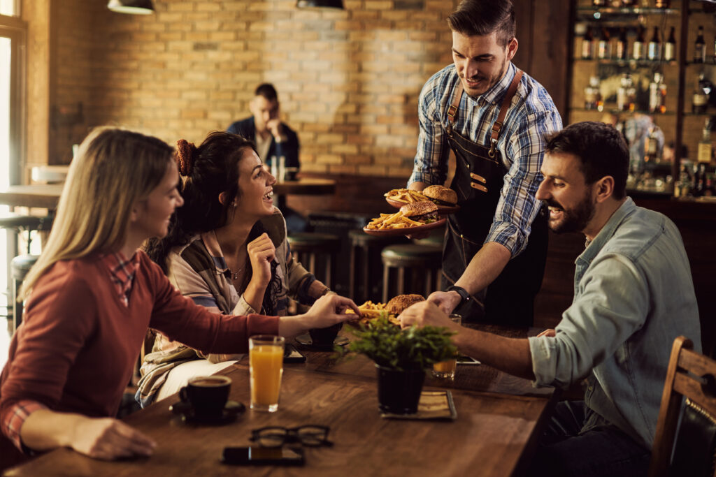 Group of happy friends having fun while waiter is serving them food in a pub.