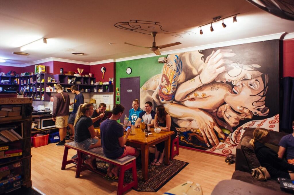 A communal dining room and kitched where travellers are sitting at the table chatting and cookin gdinner in the kitchen area at Beachside Backpackers