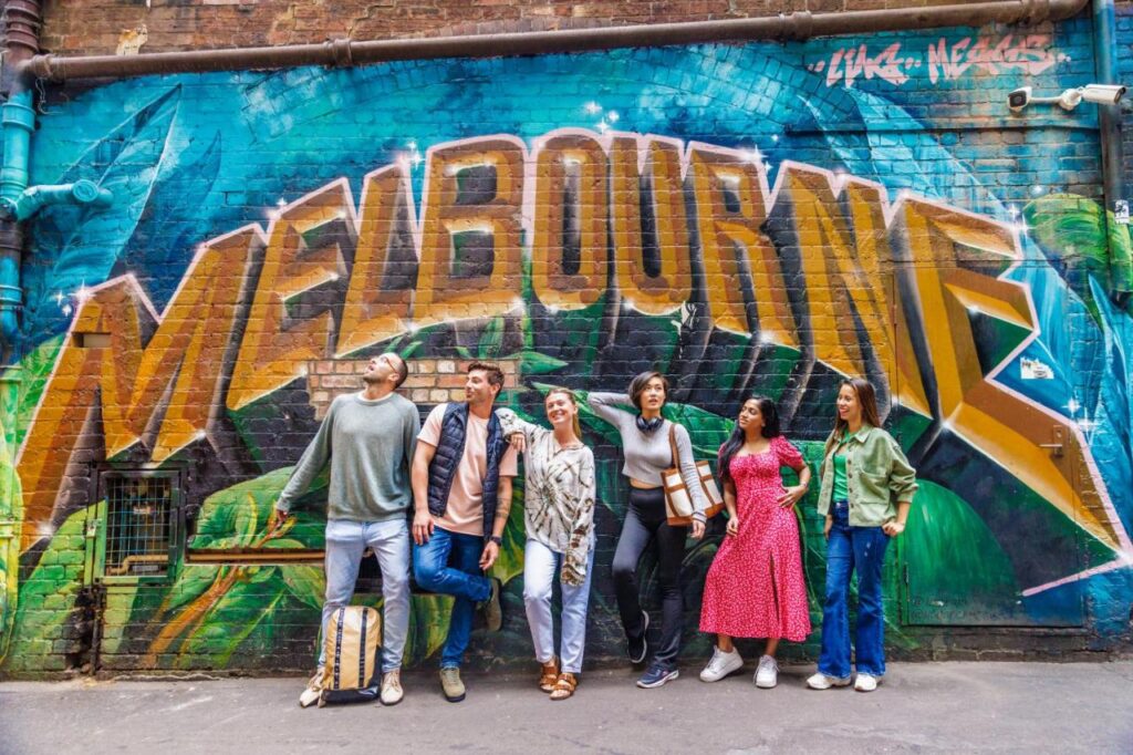 A bunch of travellers having a photo taken in front of a murial displaying the words MELBOURSE in gols and a blue background at YHA Melbourne Central