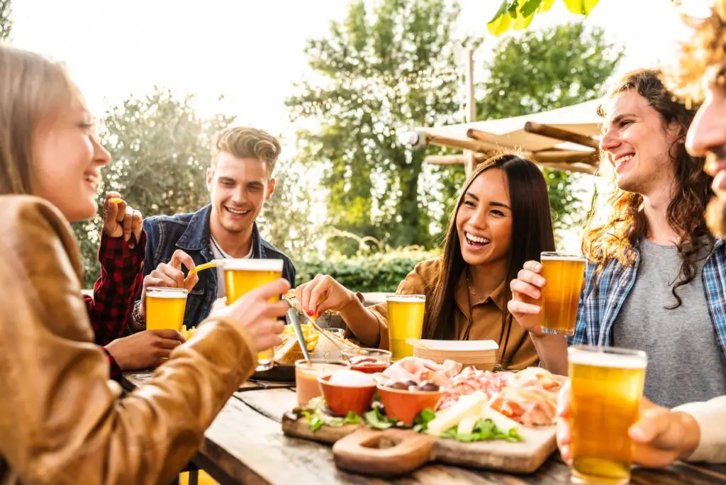 Group of multi ethnic friends having backyard dinner party together - Diverse young people sitting at bar table toasting beer glasses in brewery pub garden - Happy hour, lunch break. Bondi Beach New South Wales